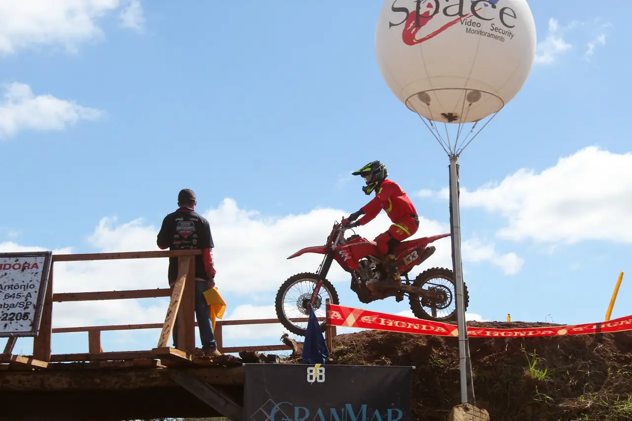 Piloto passando pela ponte de madeira da pista, com público e o balão de um patrocinador ao fundo